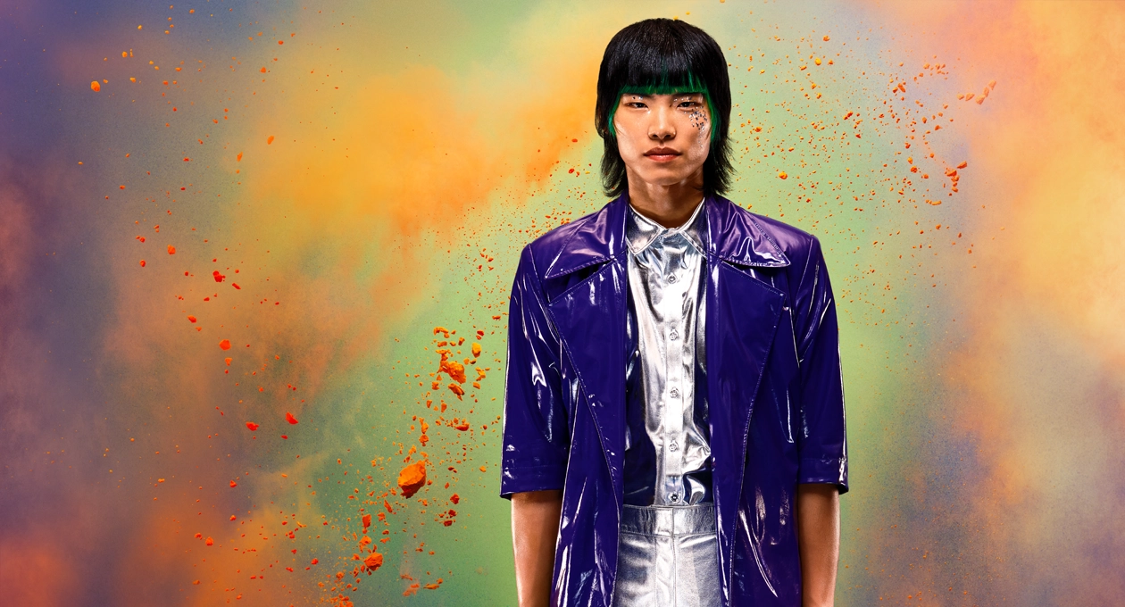A person in a silver shirt and shiny purple jacket stands against a vibrant multicolored background, showcasing the brightness, contrast, and wide color gamut available on Studio Display XDR