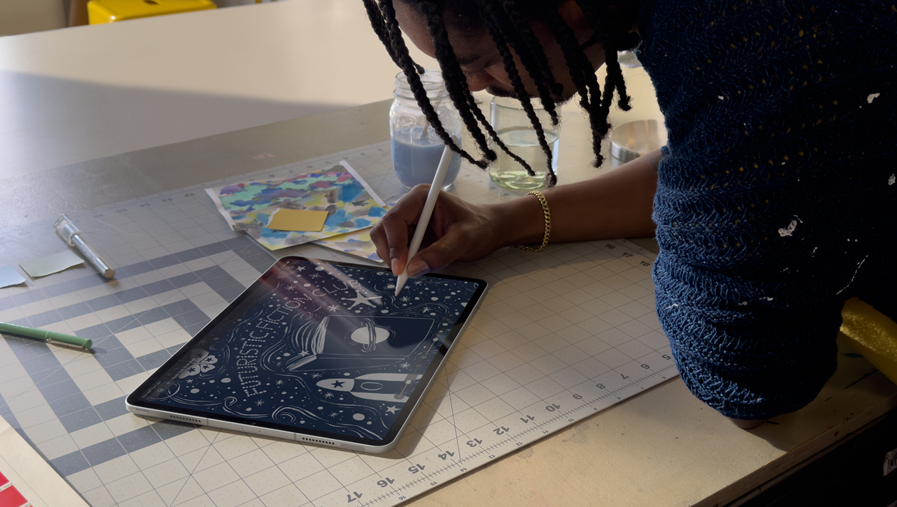 Person with braids wearing a blue sweater drawing on iPad Pro with Apple Pencil, screen shows illustration of a night sky with a planet on a book cover, space ship, butterfly and futuristic fiction club written in the centre