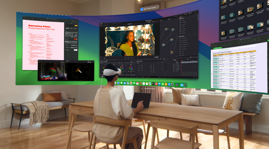 A person wearing Apple Vision Pro sits at a desk with a 13-inch MacBook Pro open, and a curved, ultra-wide display of their screen contents projects into the room