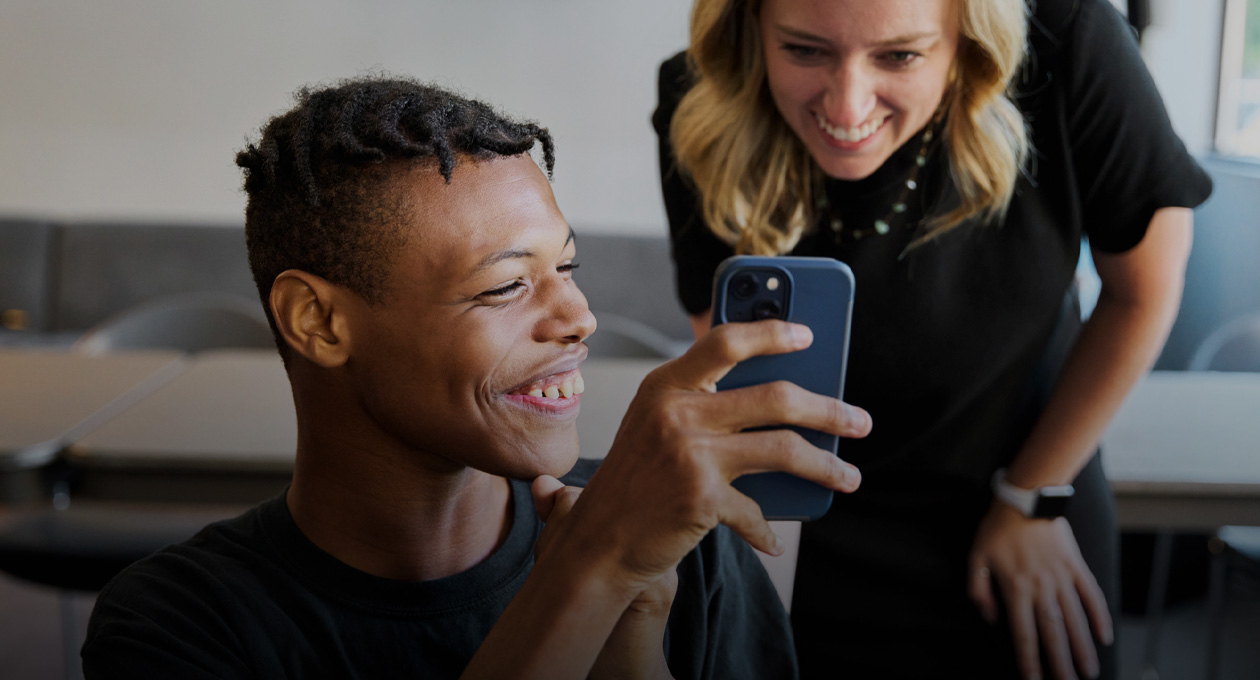 Photo of a young man with low vision smiling and showing his iPhone screen to a smiling woman standing behind his left shoulder