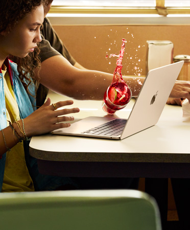 En person strekker seg etter noe på en restaurant og søler en kald drikke over en åpen MacBook.