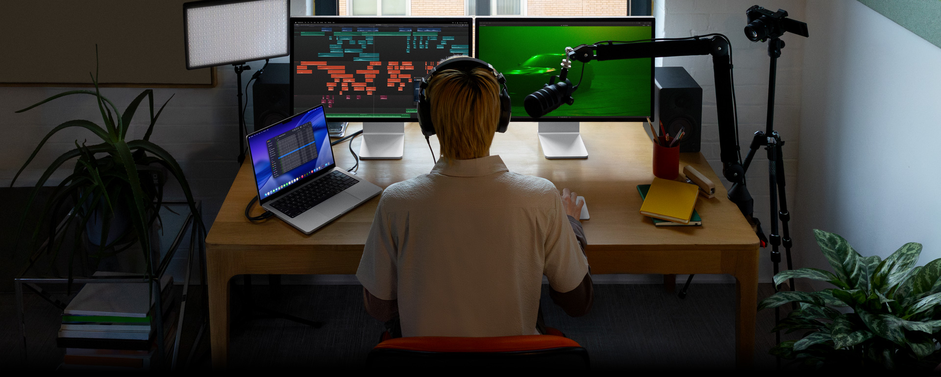 Person sitting at desk with mic and headphones in front of three devices, Mac Studio Display showing detailed audio editing tools, sound clips in different blocks of color, a second Mac Studio Display screen showing car with moody green lighting, MacBook Pro 16 screen long list of shots in a Final Cut Pro project