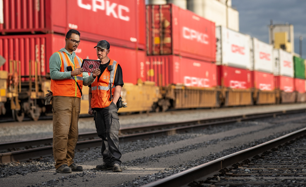 Two railroad workers reference an iPad while standing in a train yard beside a stationary train carrying shipping containers
