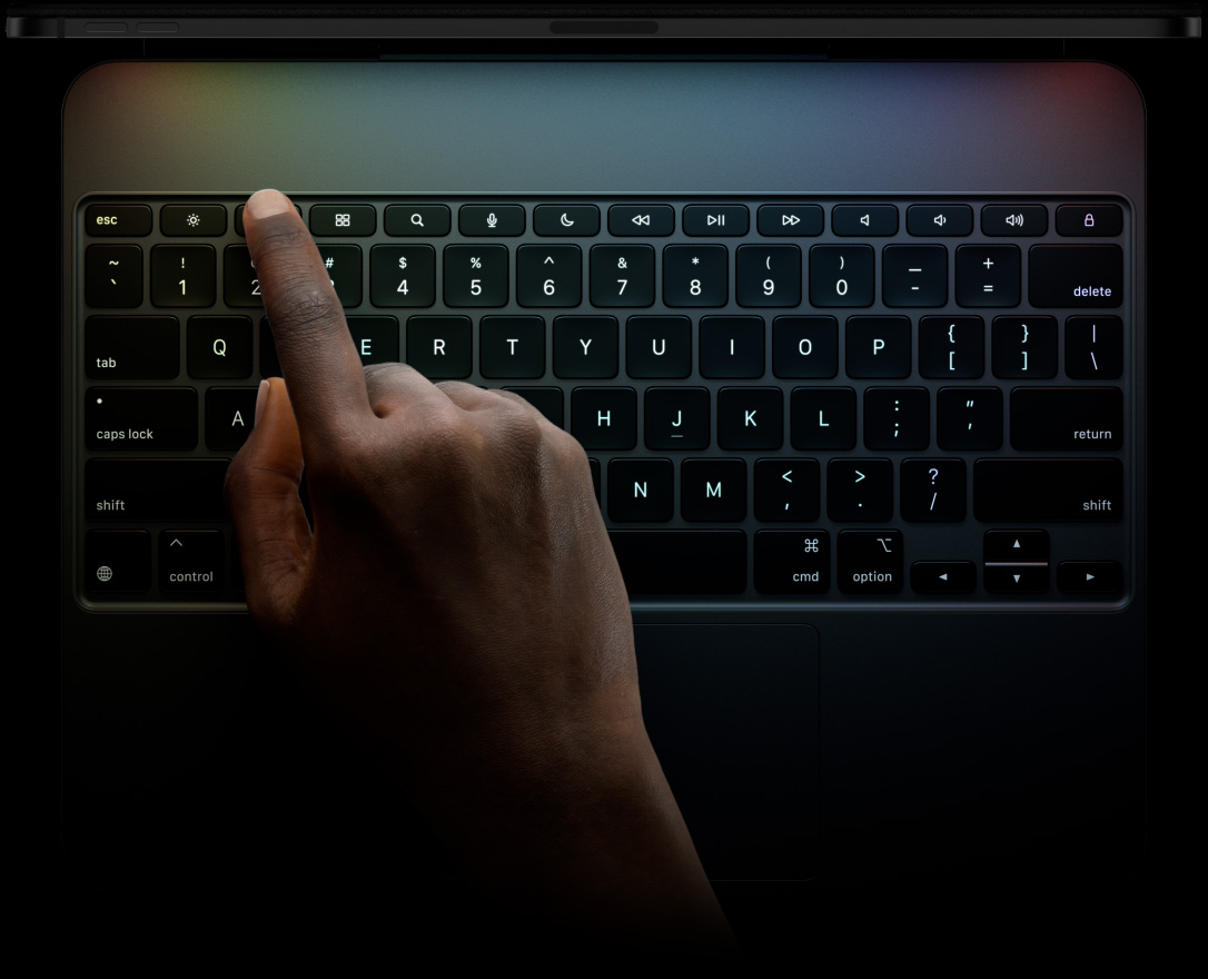 Imagem da borda superior do iPad Pro preto-espacial na horizontal conectado a um Magic Keyboard. Imagem da parte da frente na horizontal com teclas pretas retroiluminadas, teclas de função dedicadas, escape, tab, caps lock, short, control, option, shift, delete, return, teclas de direção com disposição em “T” invertido e trackpad integrado