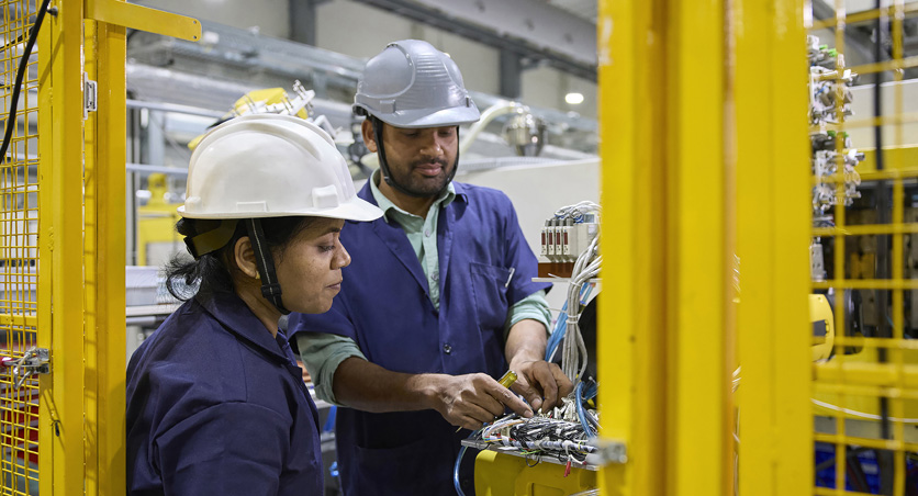 Two employees in work smocks and safety hats stand in front of an assembly machine
