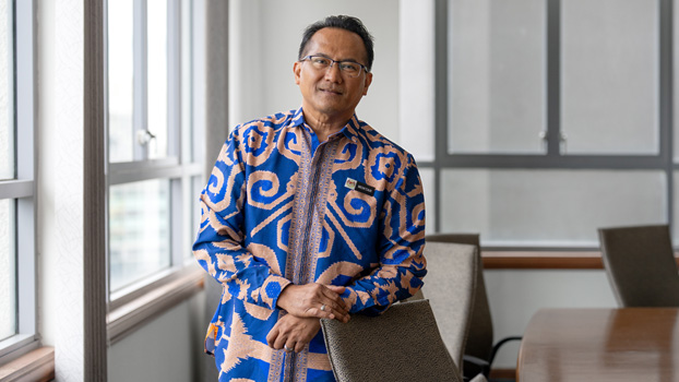 Moktar Wahid smiling while sitting at a school desk with a closed MacBook and iPad next to him.